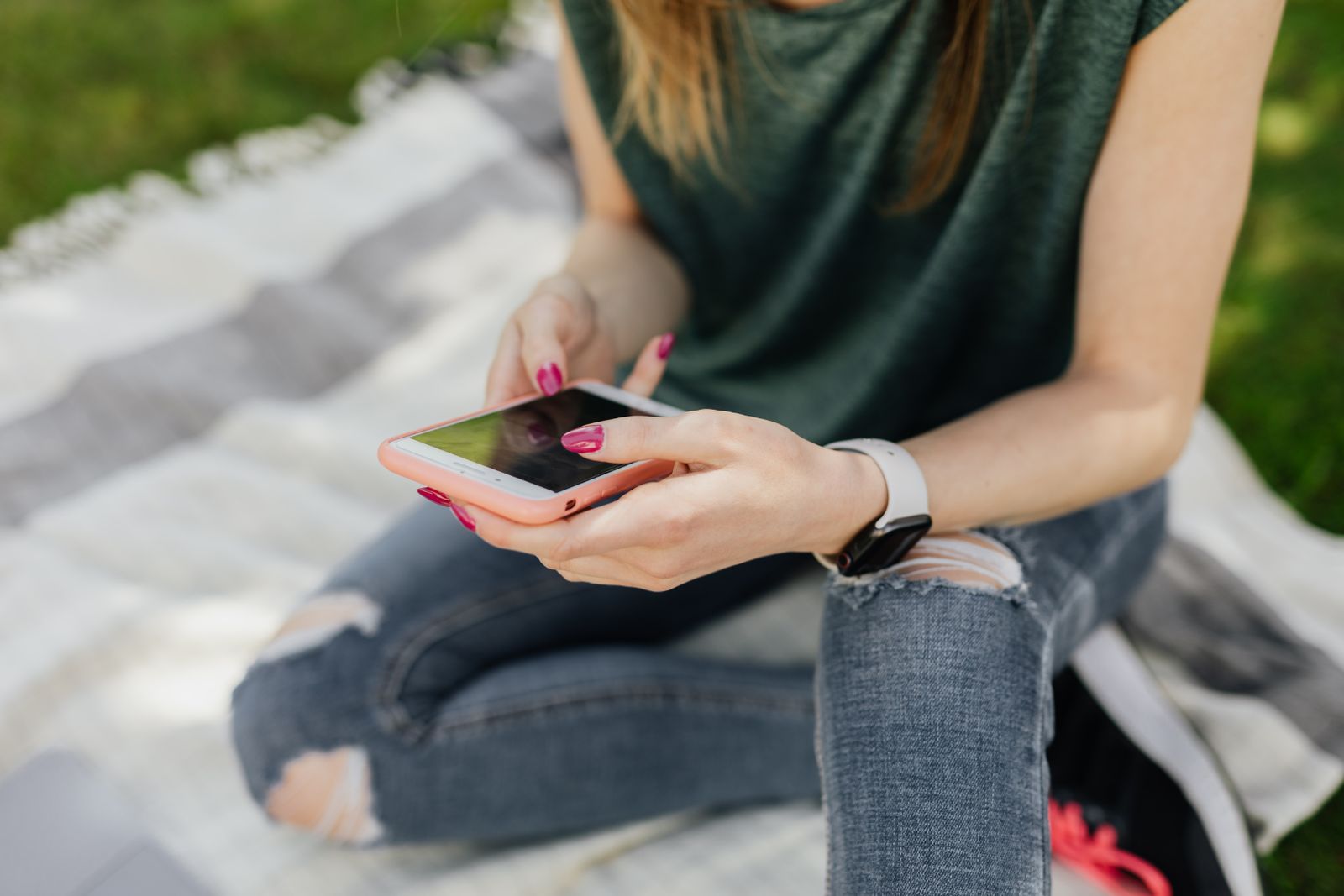 Crop faceless slim female in casual outfit surfing social media on smartphone while enjoying spare time sitting on blanket placed on green lawn in sunny park