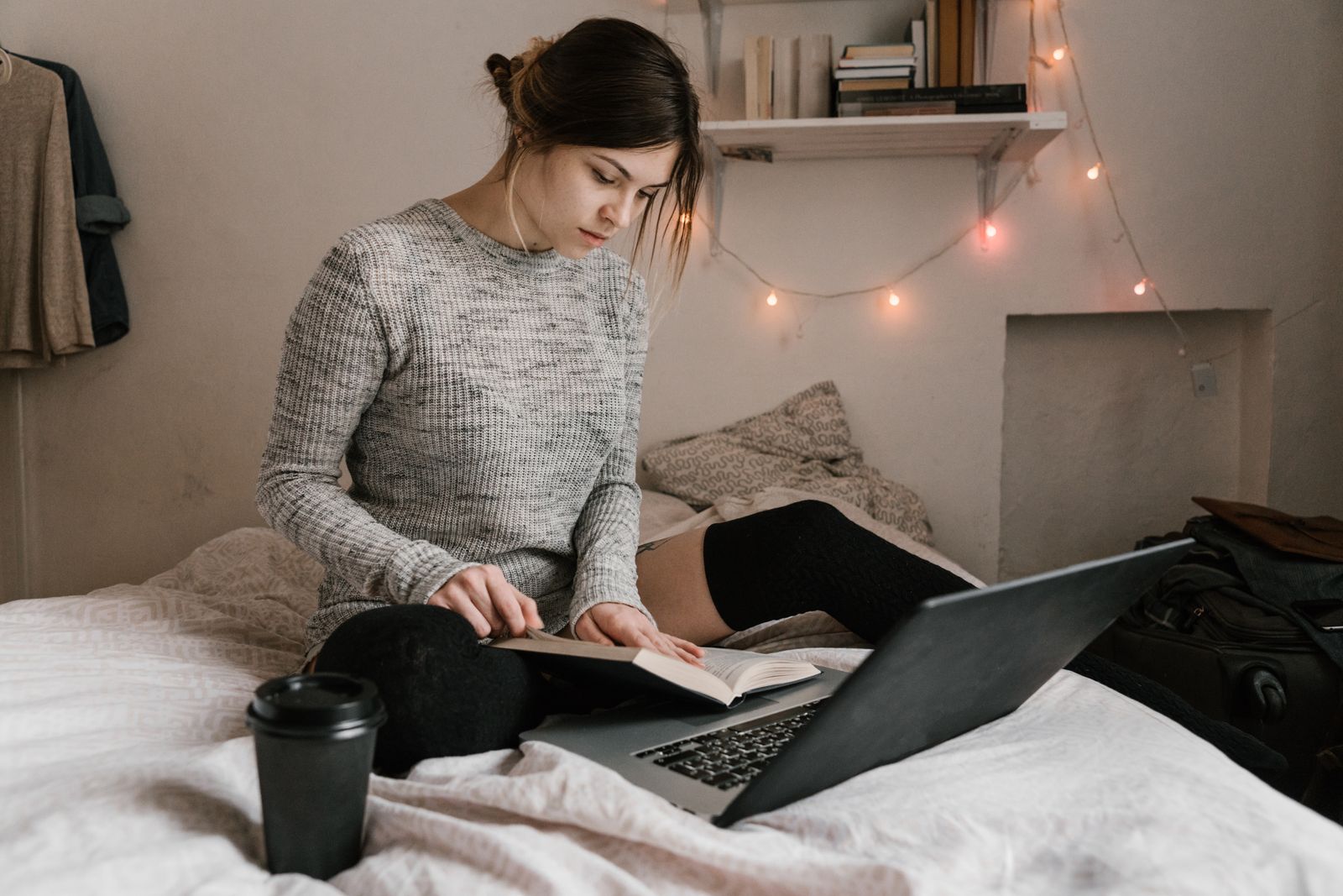 Young woman reading and studying on bed with a laptop in a cozy bedroom setting.