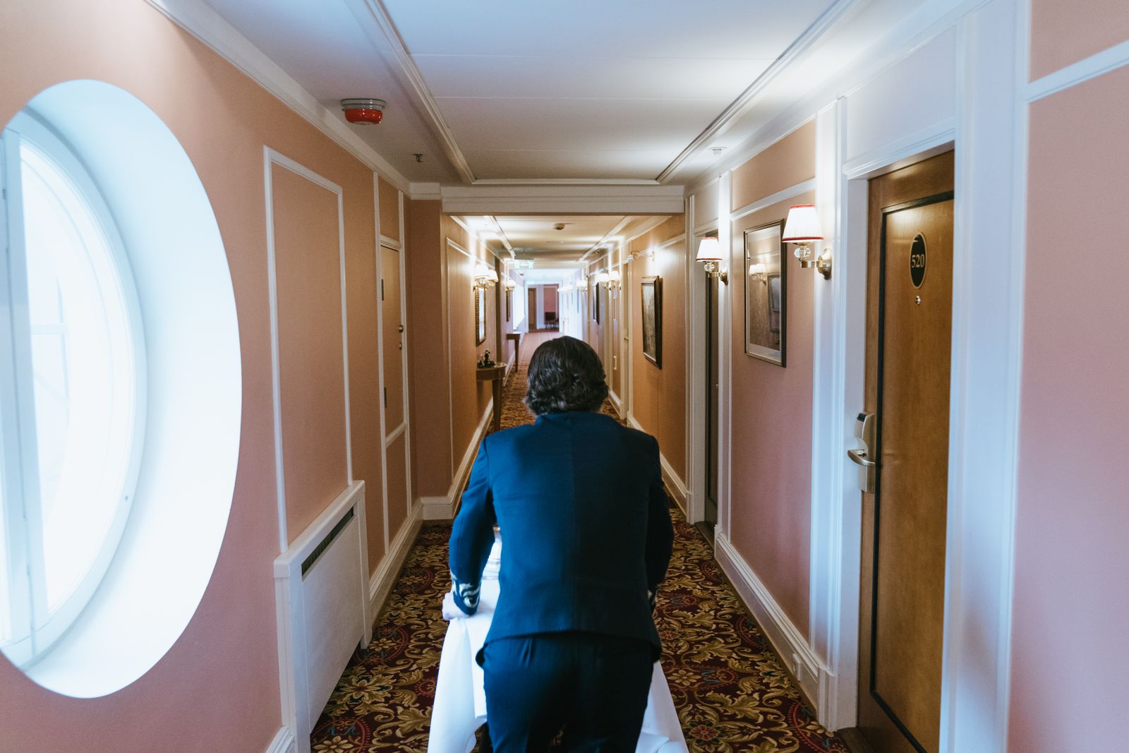 A hotel staff member in a suit pushing a cart down a well-lit corridor.