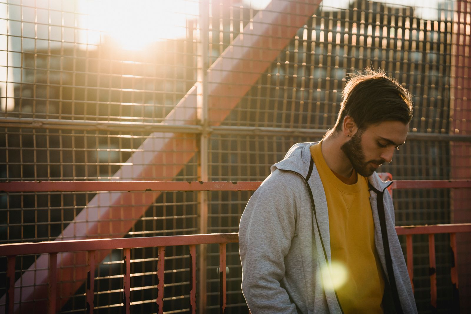 Young upset unshaven male in casual apparel looking down near grid fence in back lit in town