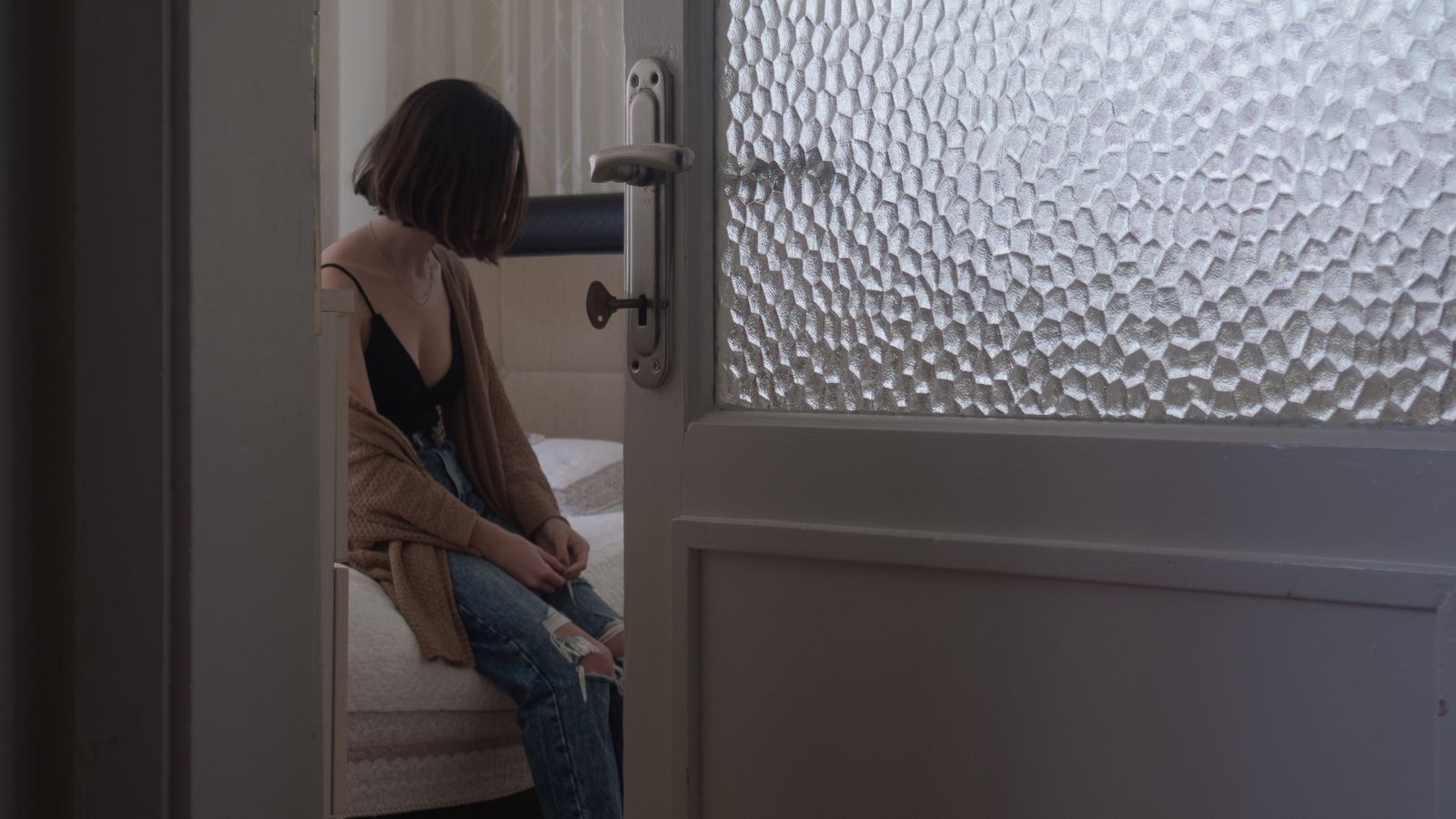 A woman sits thoughtfully on a bed in a dimly lit bedroom, evoking feelings of solitude and reflection.