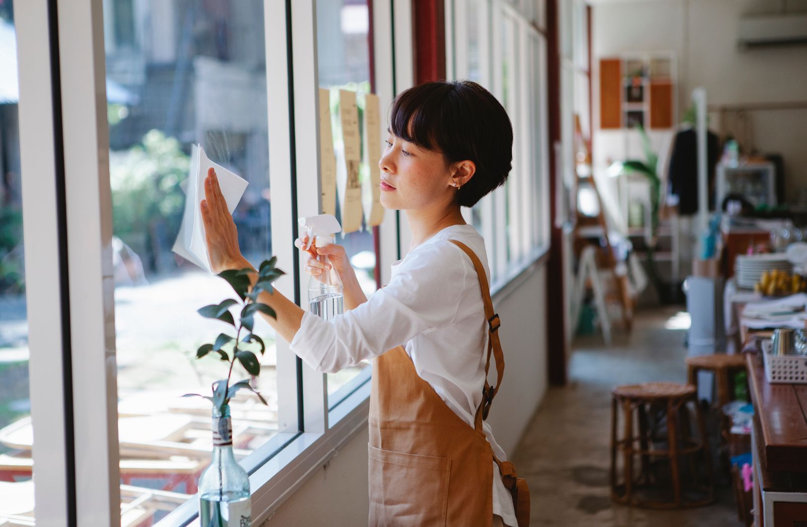 Asian woman cleaning a restaurant window with a spray bottle and tissue.