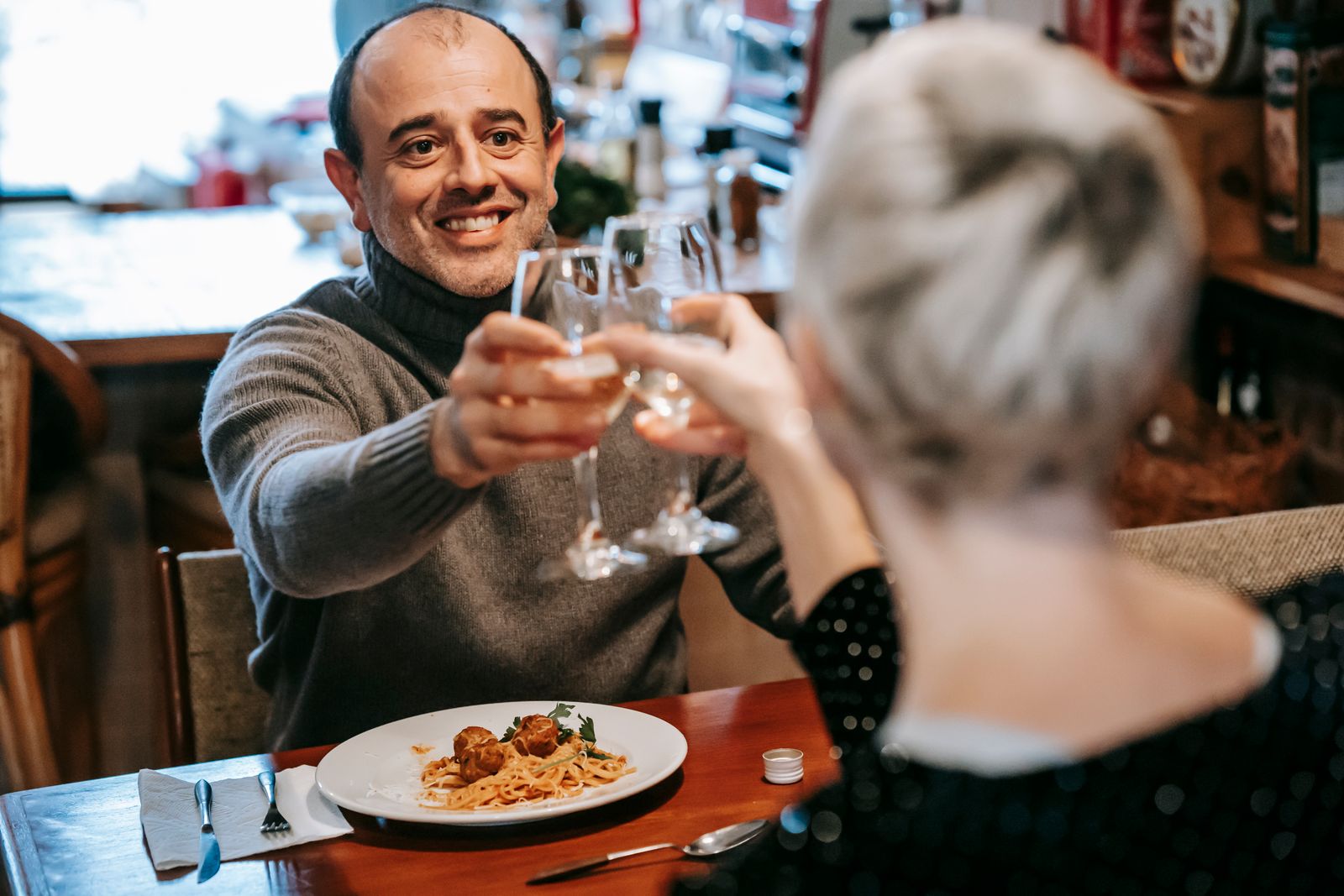 Smiling couple sitting in light room while having dinner with pasta with meatballs on plate near cutlery and clinking glasses with wine while looking at each other
