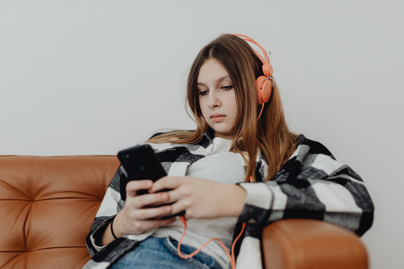 Teenage girl sitting on a sofa listening to music on headphones and using a smartphone.