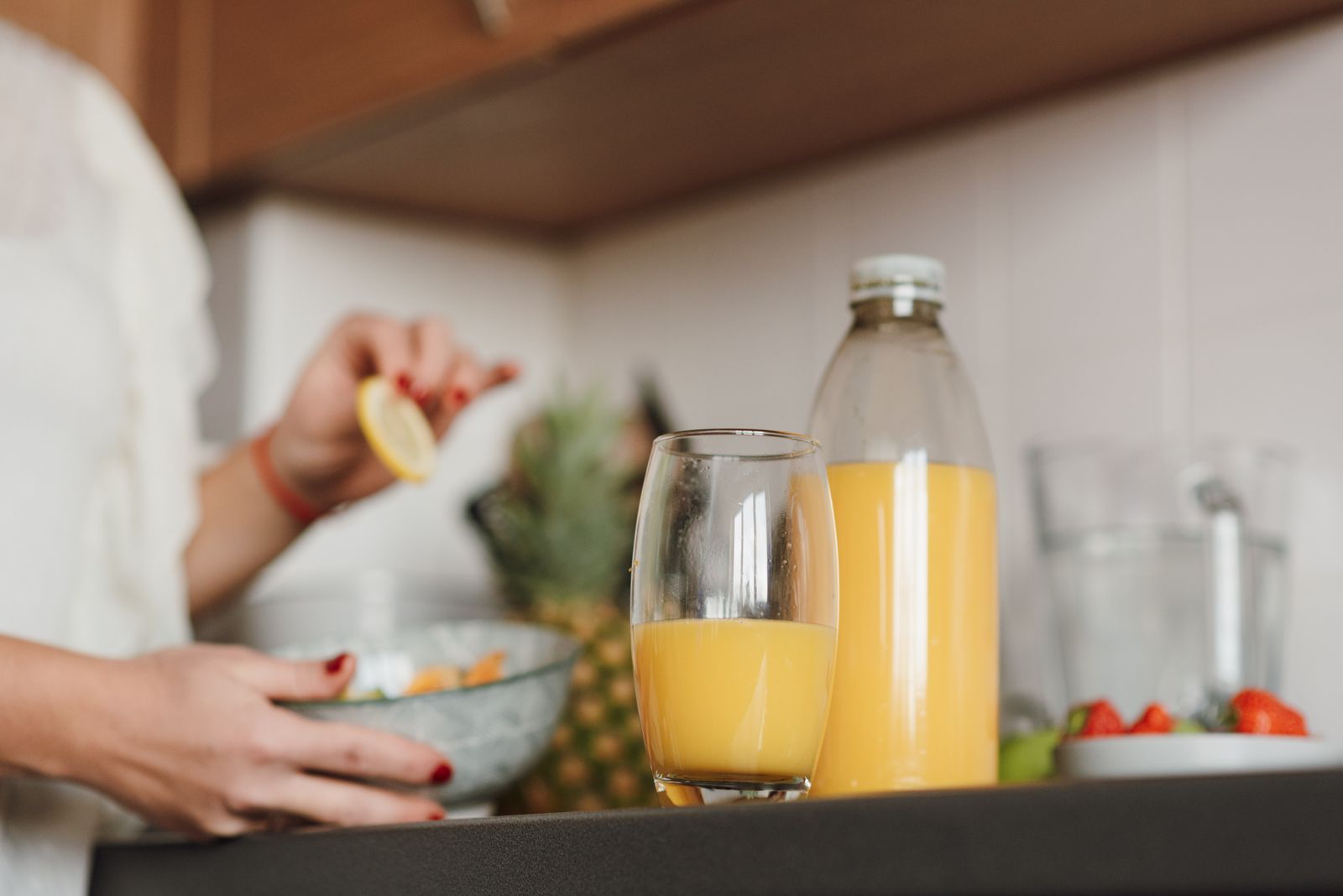 Crop unrecognizable female standing near table with fresh orange juice while cooking fruit salad in kitchen