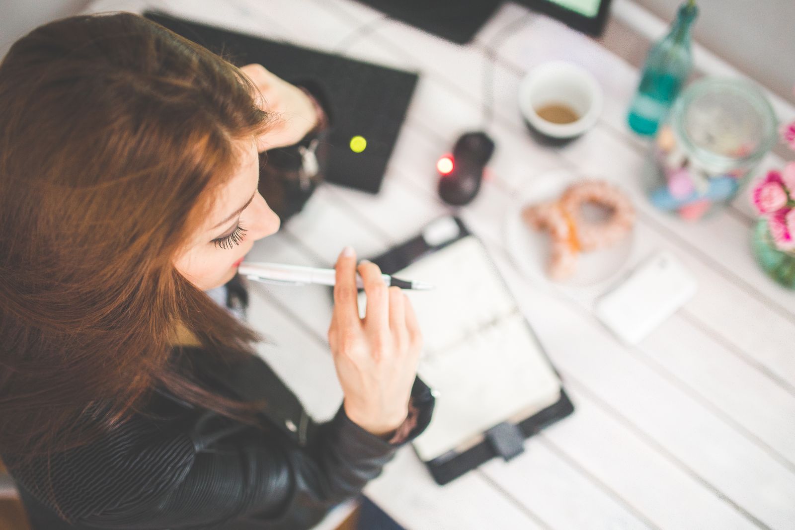 Overhead view of a young woman studying at a home office desk, thinking and taking notes.