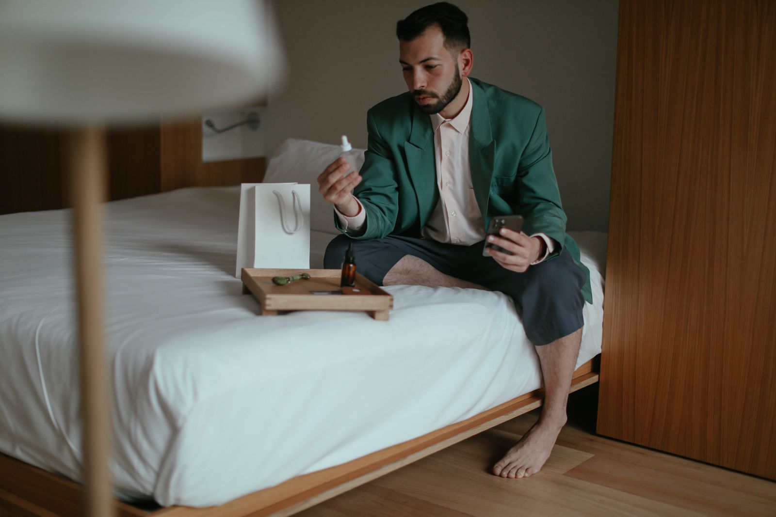 Adult man sitting on bed examining skincare product, emphasizing relaxation and personal care.