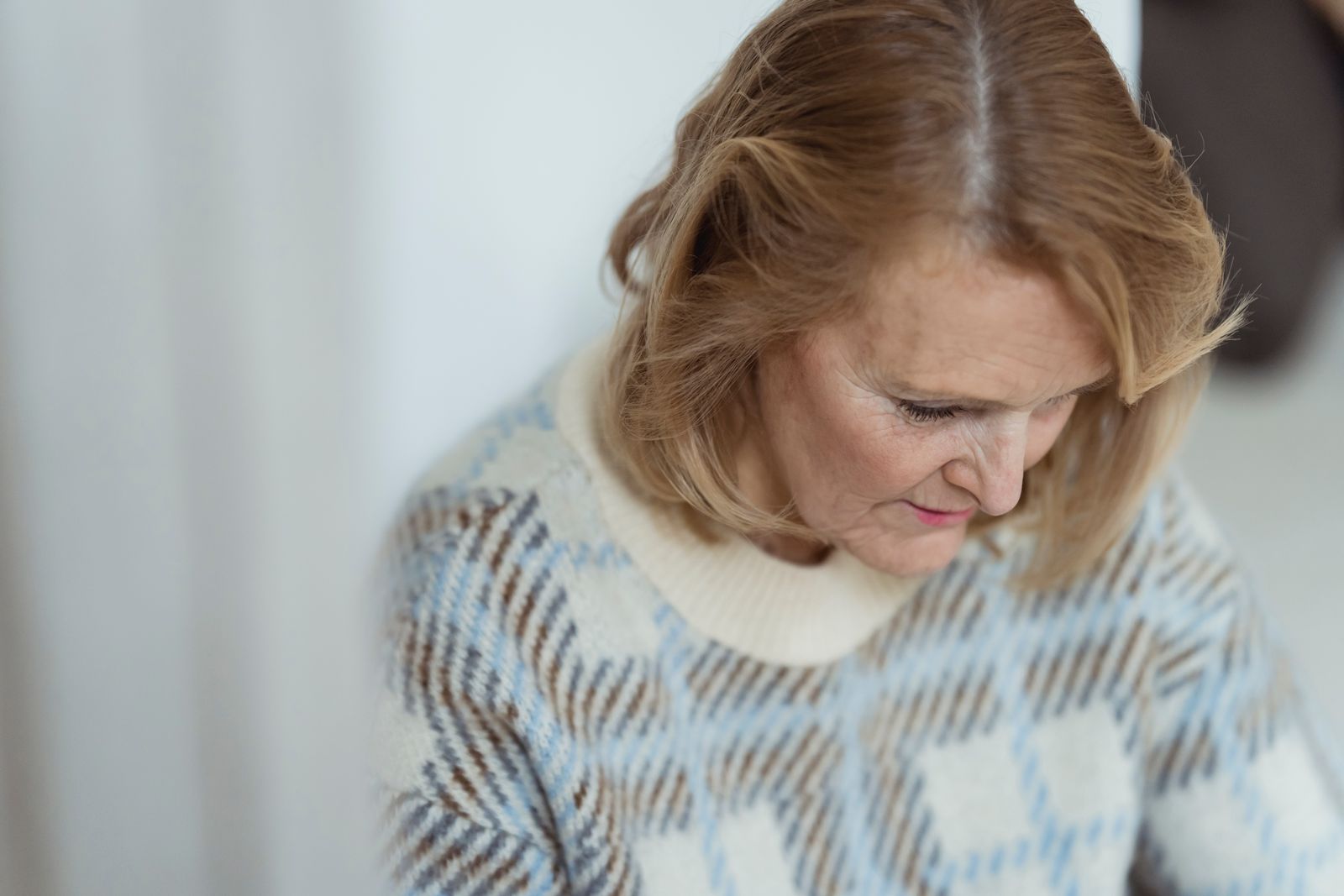 Crop positive middle aged lady with blond hair in stylish sweater smiling and looking down in daylight