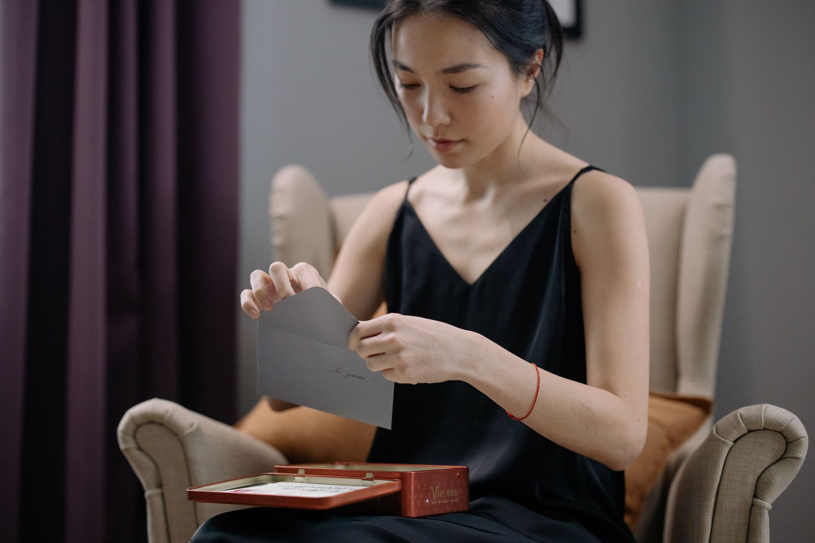 Asian woman opening a letter while sitting on an armchair in a cozy indoor setting.