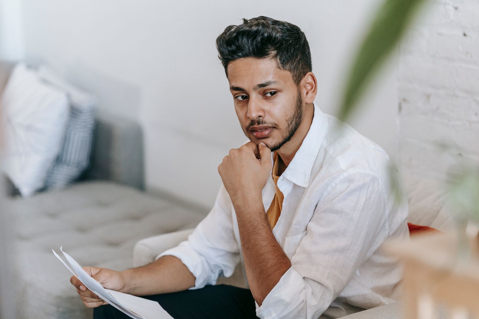 Contemplative young ethnic male in casual clothes reading documents and touching chin while sitting on couch and looking away pensively