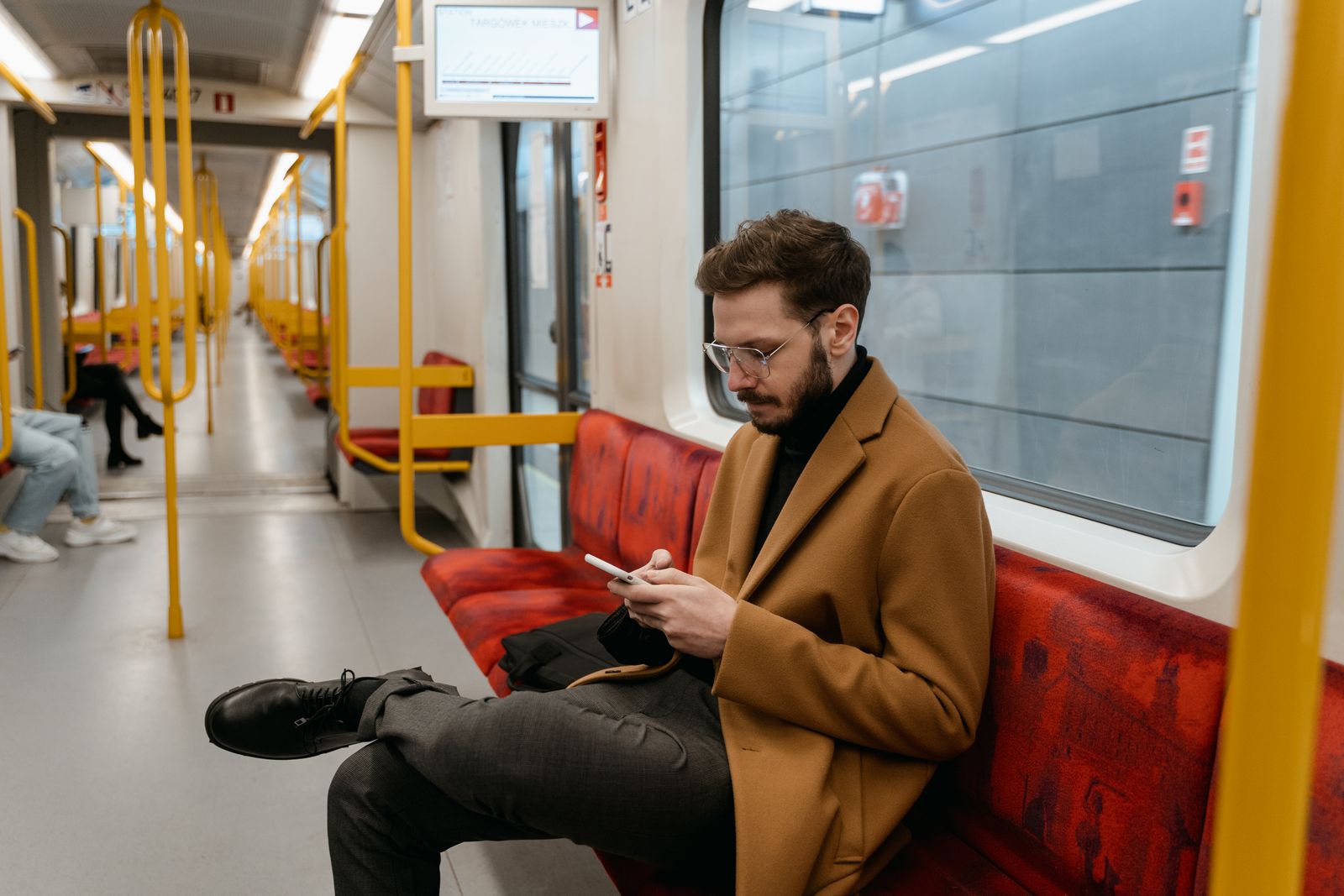 Bearded man in brown coat texting on a train with red seats. Commuting lifestyle scene.