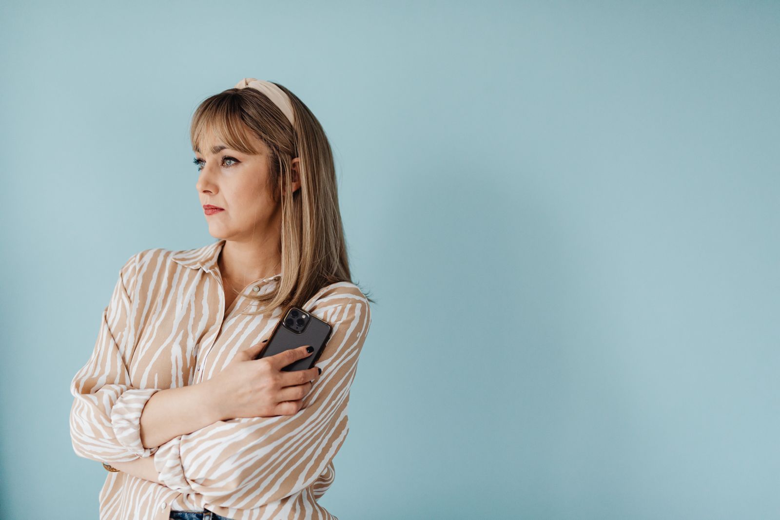 Thoughtful woman in striped shirt holding a smartphone against a light blue background.