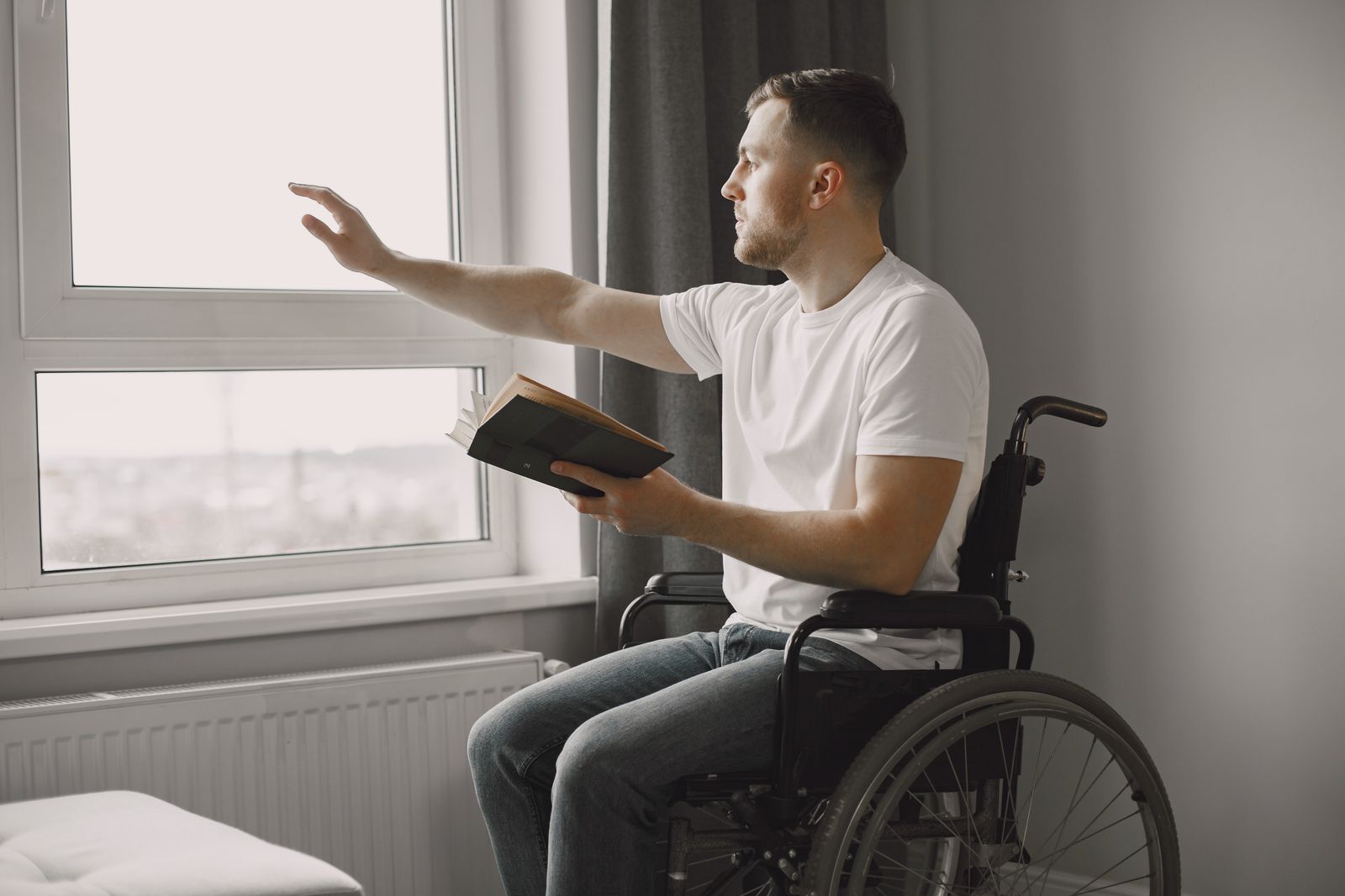 Man in a wheelchair looking through a window while holding a book indoors.