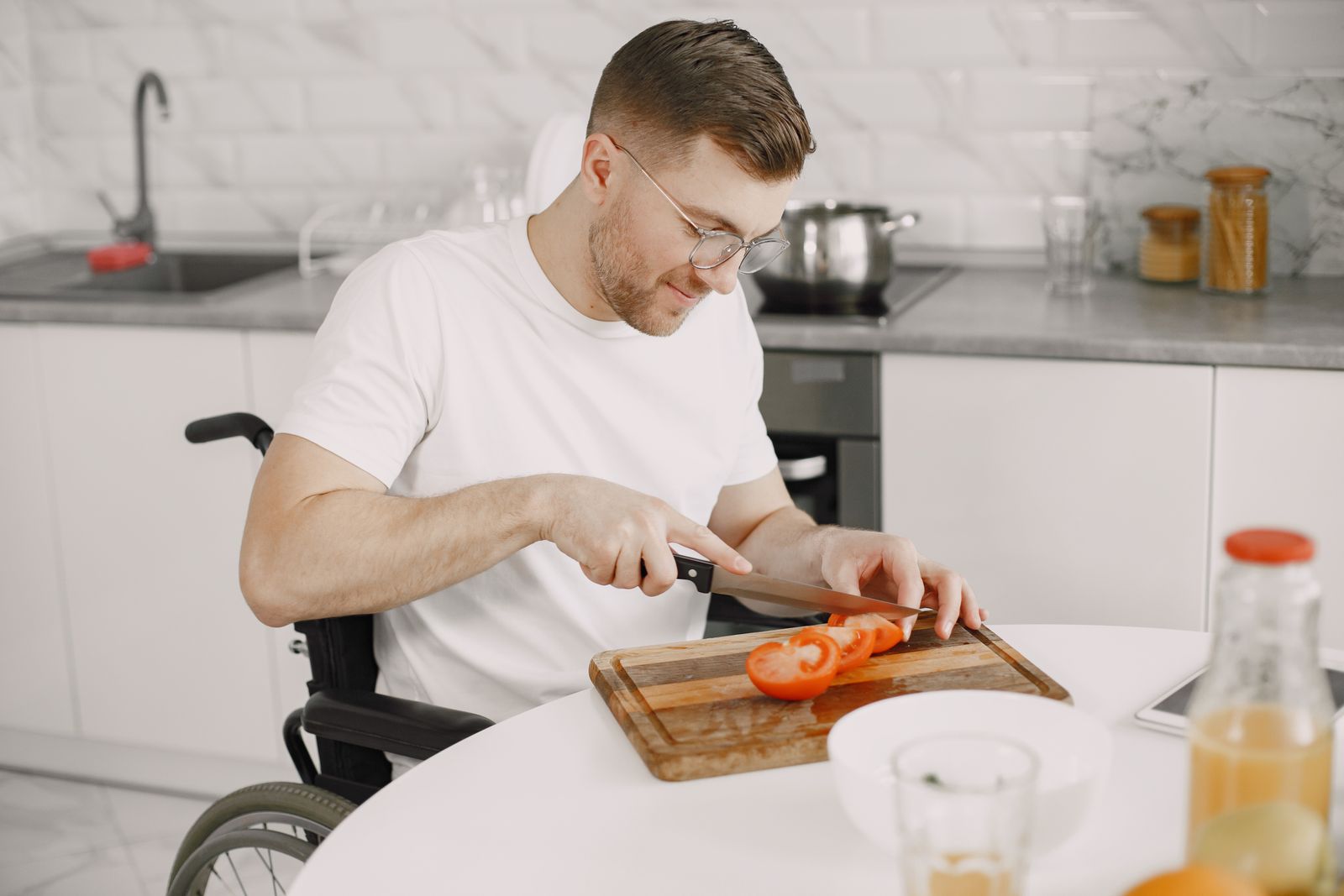 Adult man in wheelchair slicing tomato in modern kitchen setting.