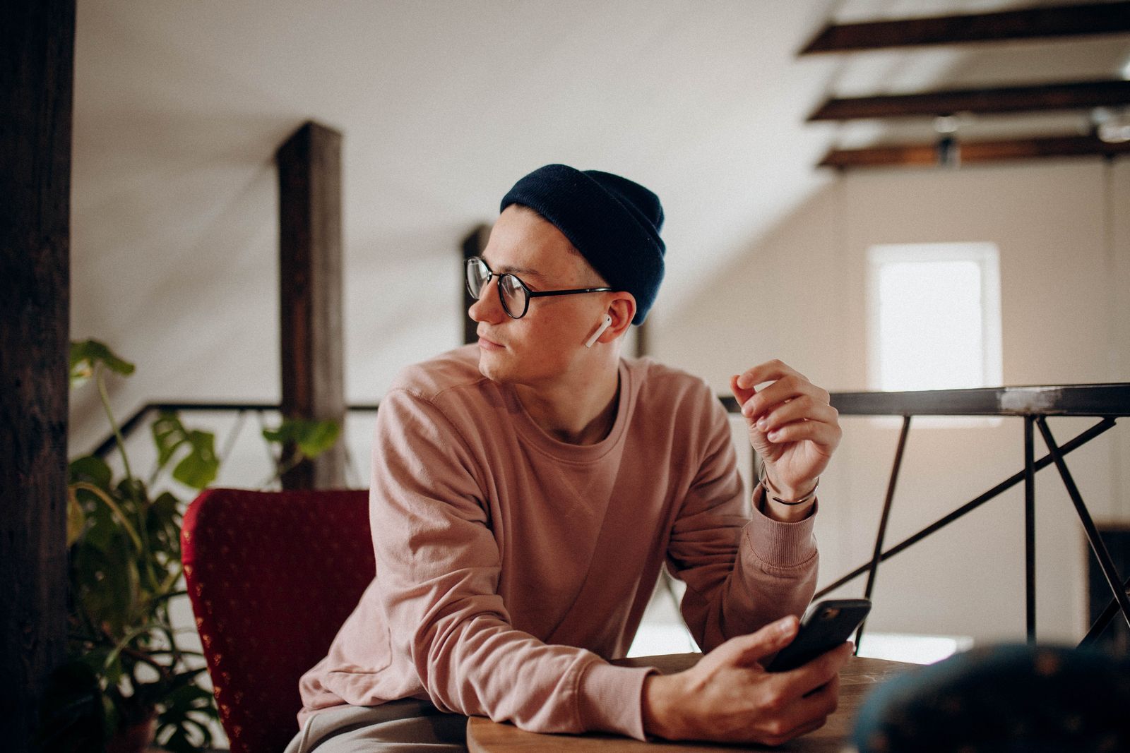 A man wearing a beanie and glasses sits indoors holding a smartphone.