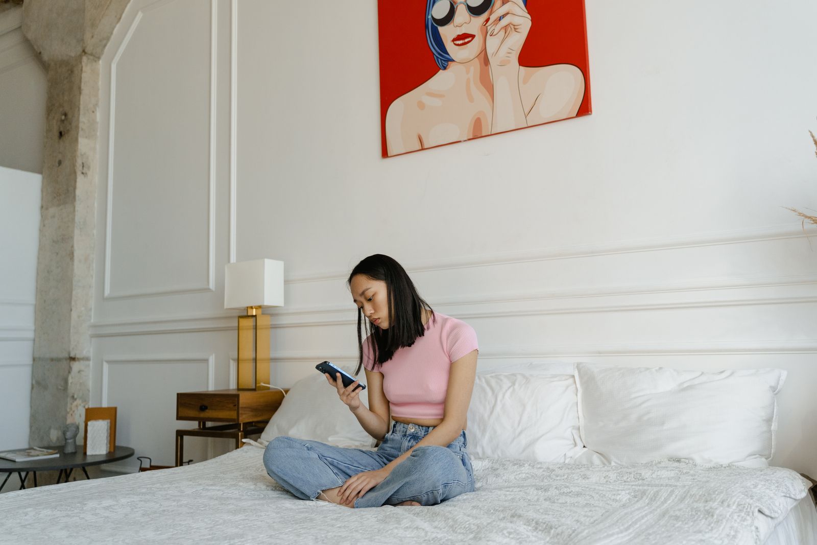 Asian woman sitting on bed using smartphone in modern bedroom with art decor.