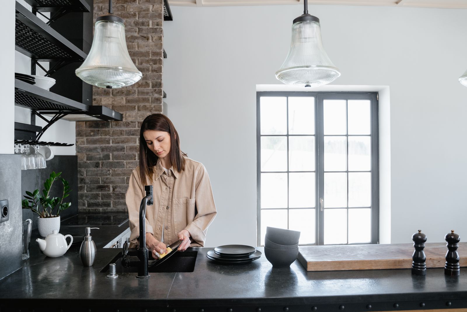 A woman washing dishes in a stylish kitchen with natural light shining through large windows.