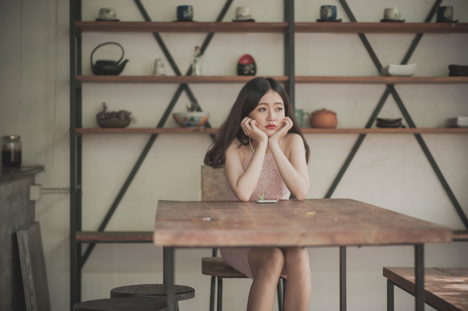 Asian woman in a pensive mood, seated at a wooden table with shelves in the background.