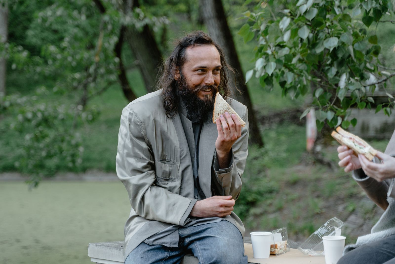 A smiling man with a beard holding a sandwich in a lush green park.