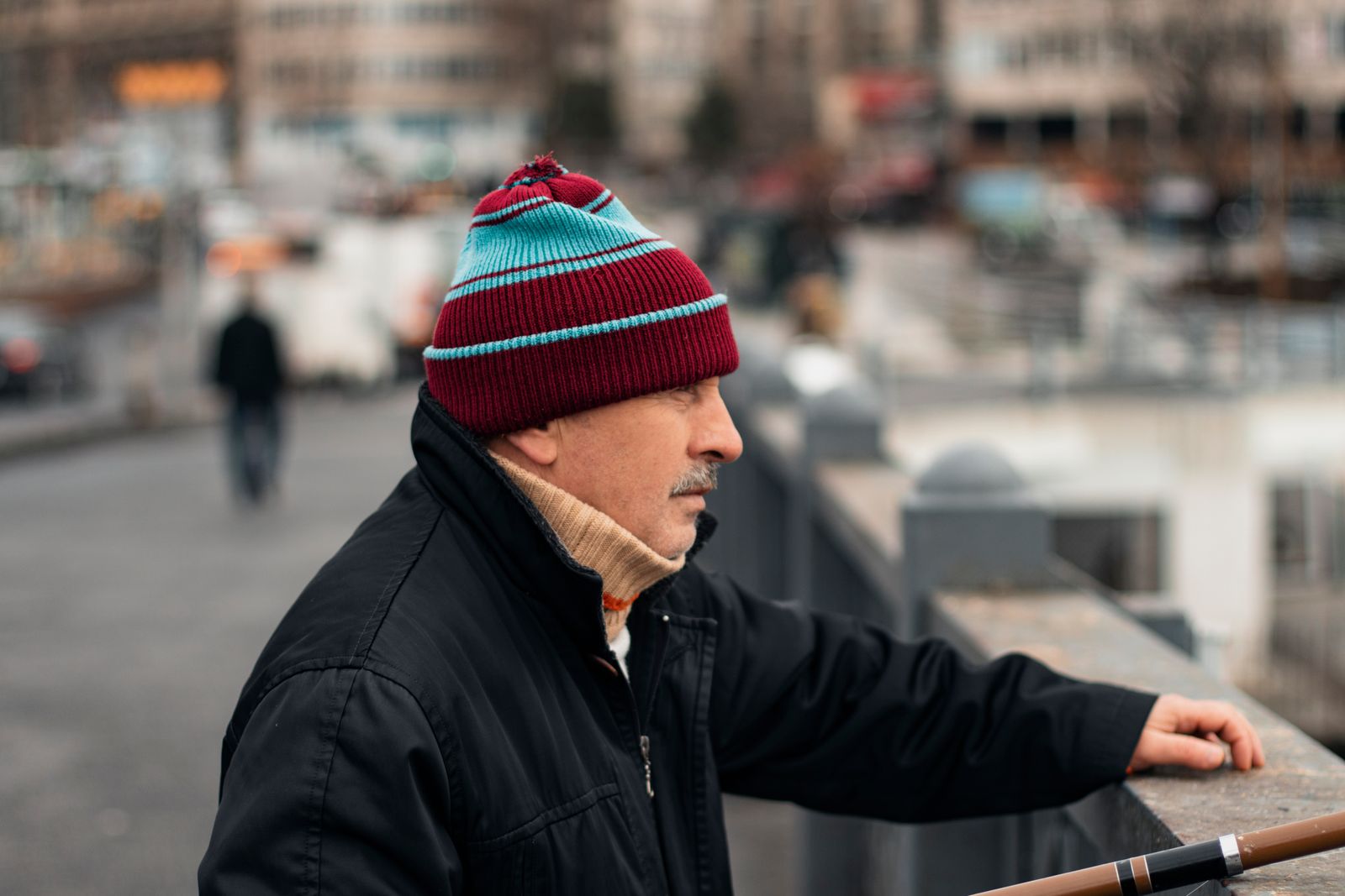 A middle-aged man wearing a colorful beanie and black jacket on a bustling bridge in Istanbul.