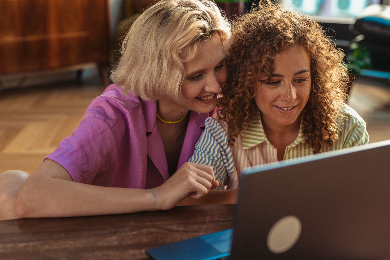 Two women smiling and working together on a laptop in a cozy indoor setting, fostering teamwork and connection.