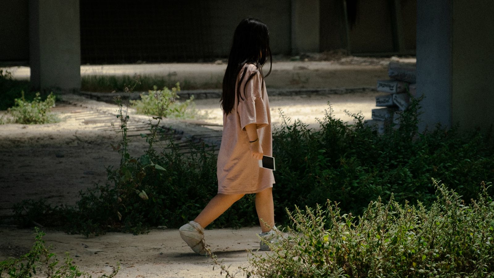 A young girl walks through a shaded urban area holding a polaroid picture.