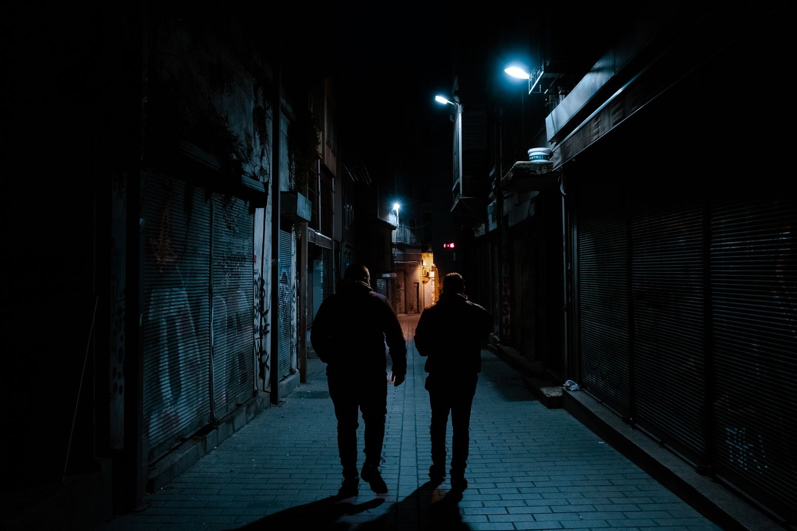 Two men silhouette walking in a dark urban alley at night with street lamps.