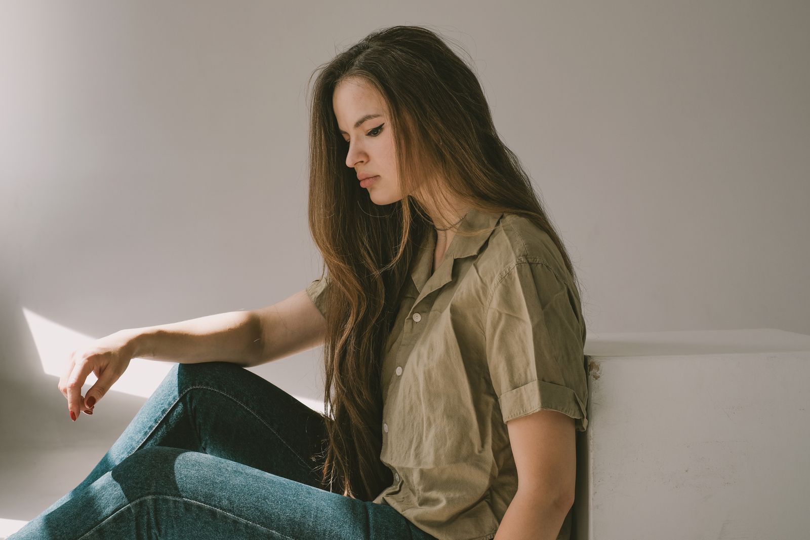 Young woman in thought sitting indoors, bathed in soft sunlight, creating a pensive mood.