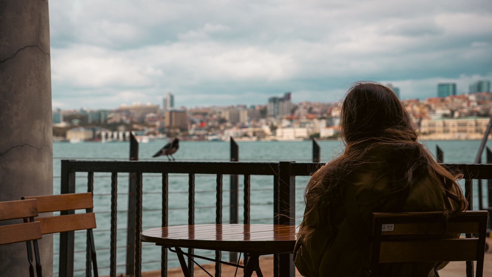A woman sits alone by the waterfront, overlooking a city skyline, creating a serene moment.