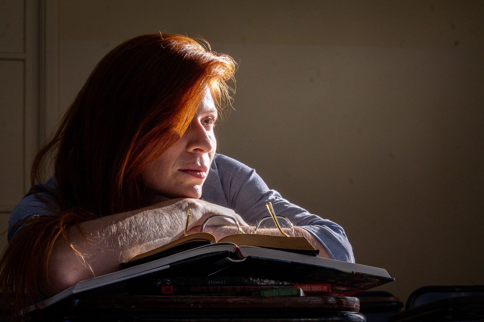 A thoughtful student sits with books in a classroom, illuminated by natural light.
