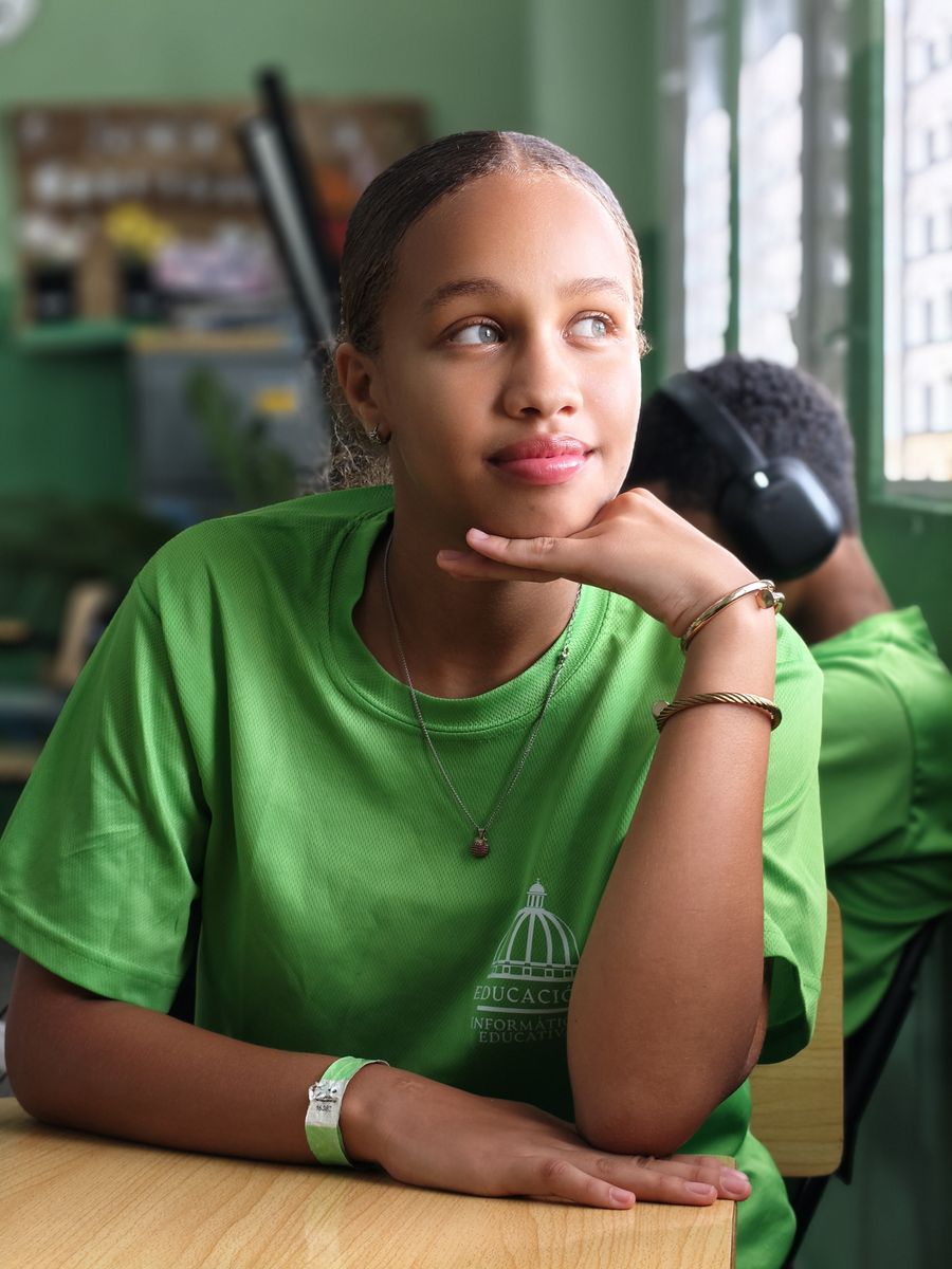 A teenage girl in a green shirt daydreams at her desk in a classroom setting.
