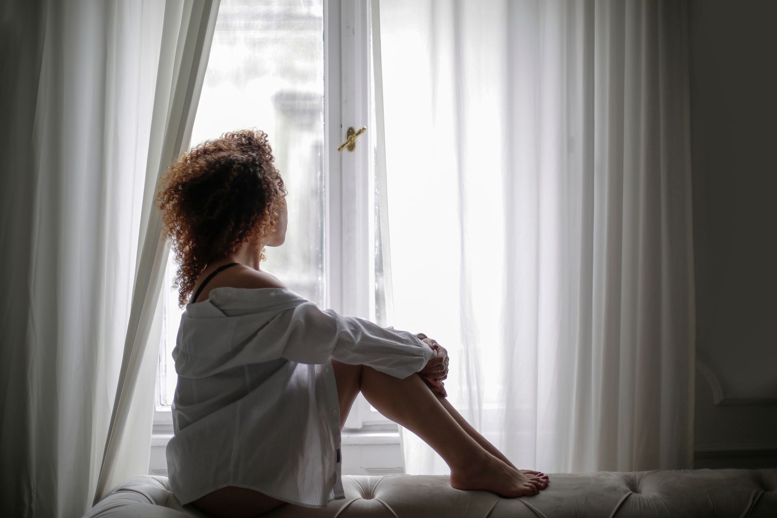 Side view of thoughtful young ethnic lady in white shirt sitting on comfortable sofa and looking through window dreamily at home