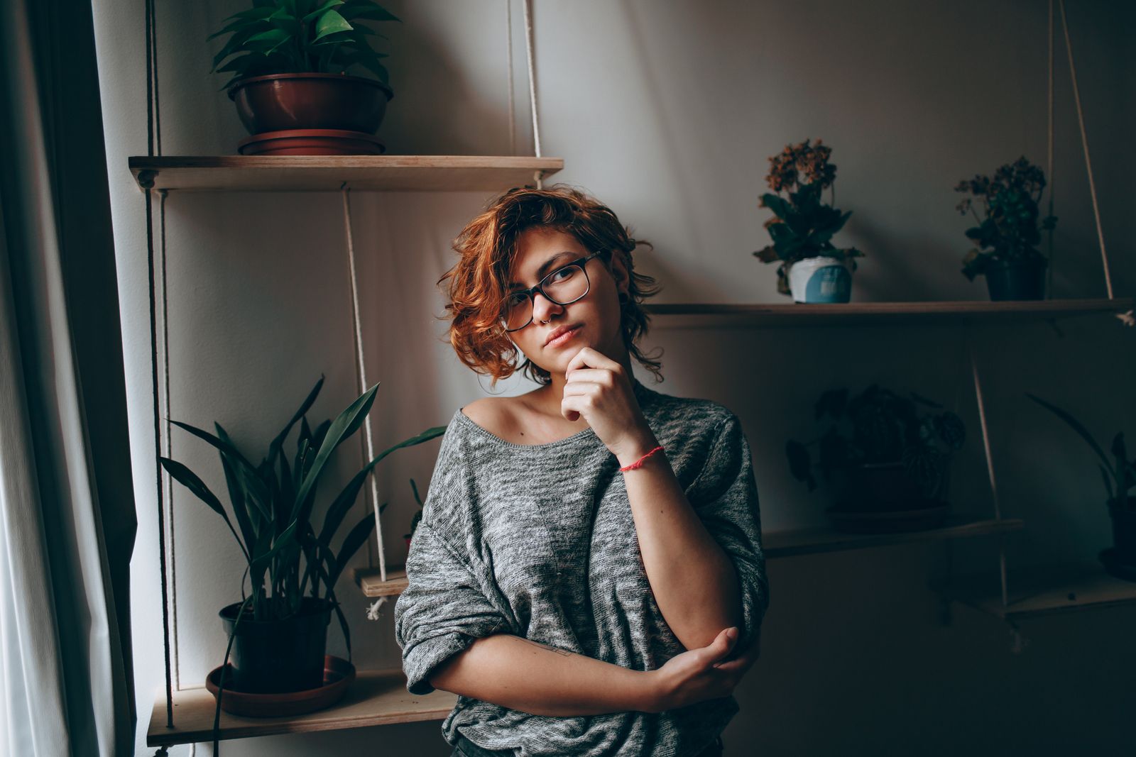 Young female in eyeglasses touching chin while thoughtfully looking at camera standing in room with plants on shelves