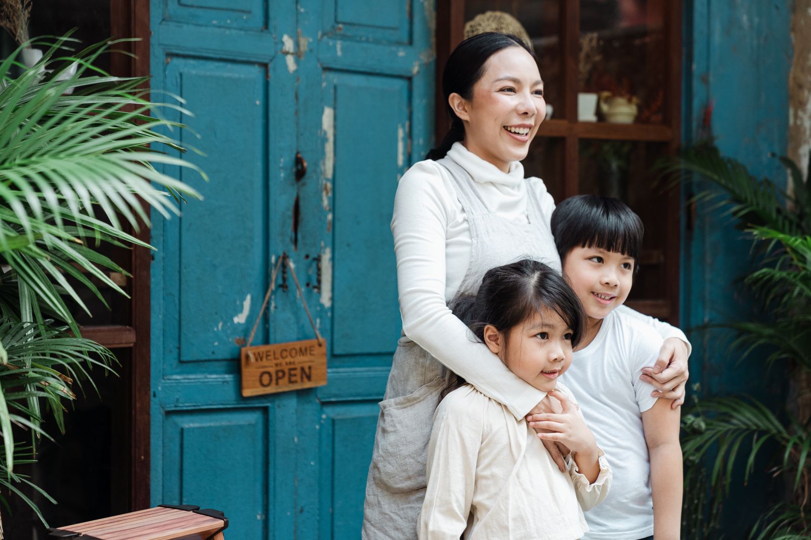 A joyful mother embraces her children outside a shop with an 'Open' sign.