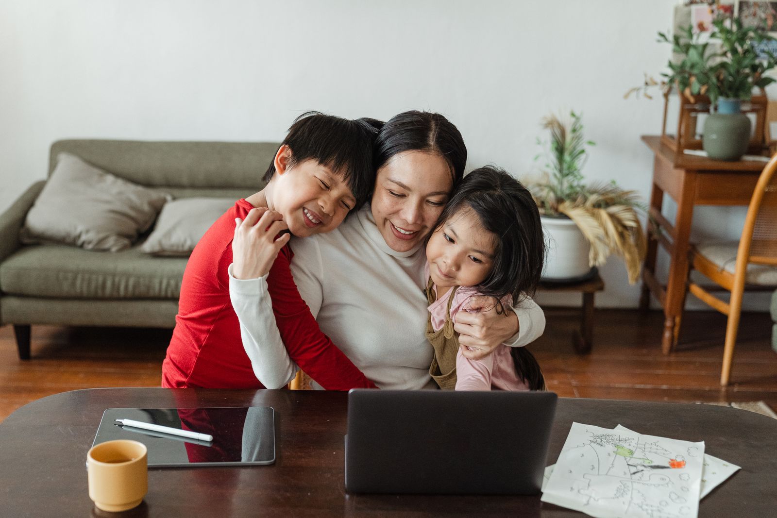 Smiling Asian mom embracing cheerful son and daughter while sitting at table with laptop and tablet and having short break from remote work