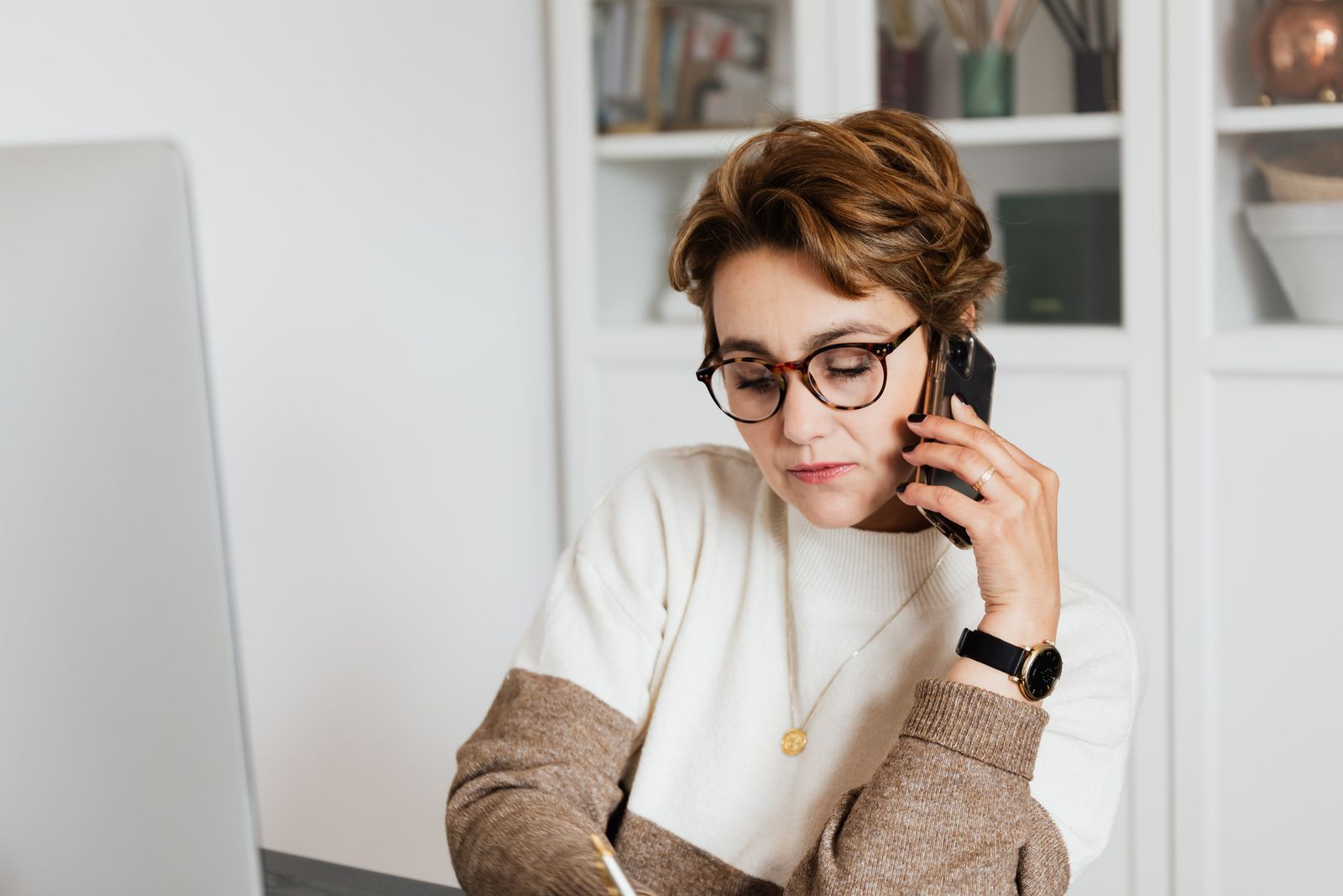 Good looking short haired female worker listening to work instructions on mobile and noting down information in modern office