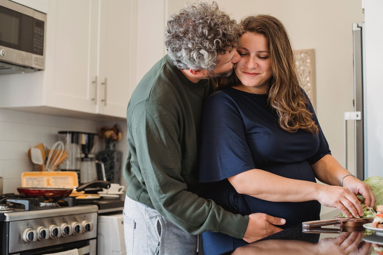 Romantic man with curly hair embracing and kissing pregnant wife cooking food in cozy kitchen