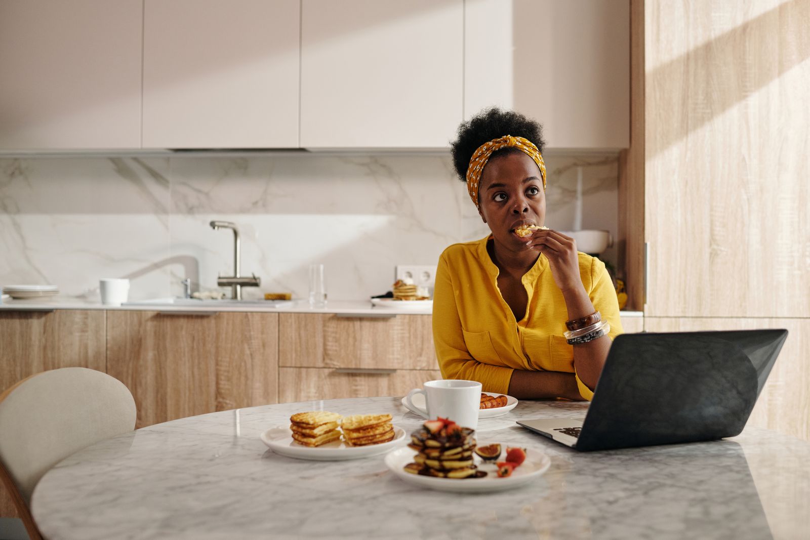 Black woman enjoying breakfast and using laptop in a modern kitchen setting.