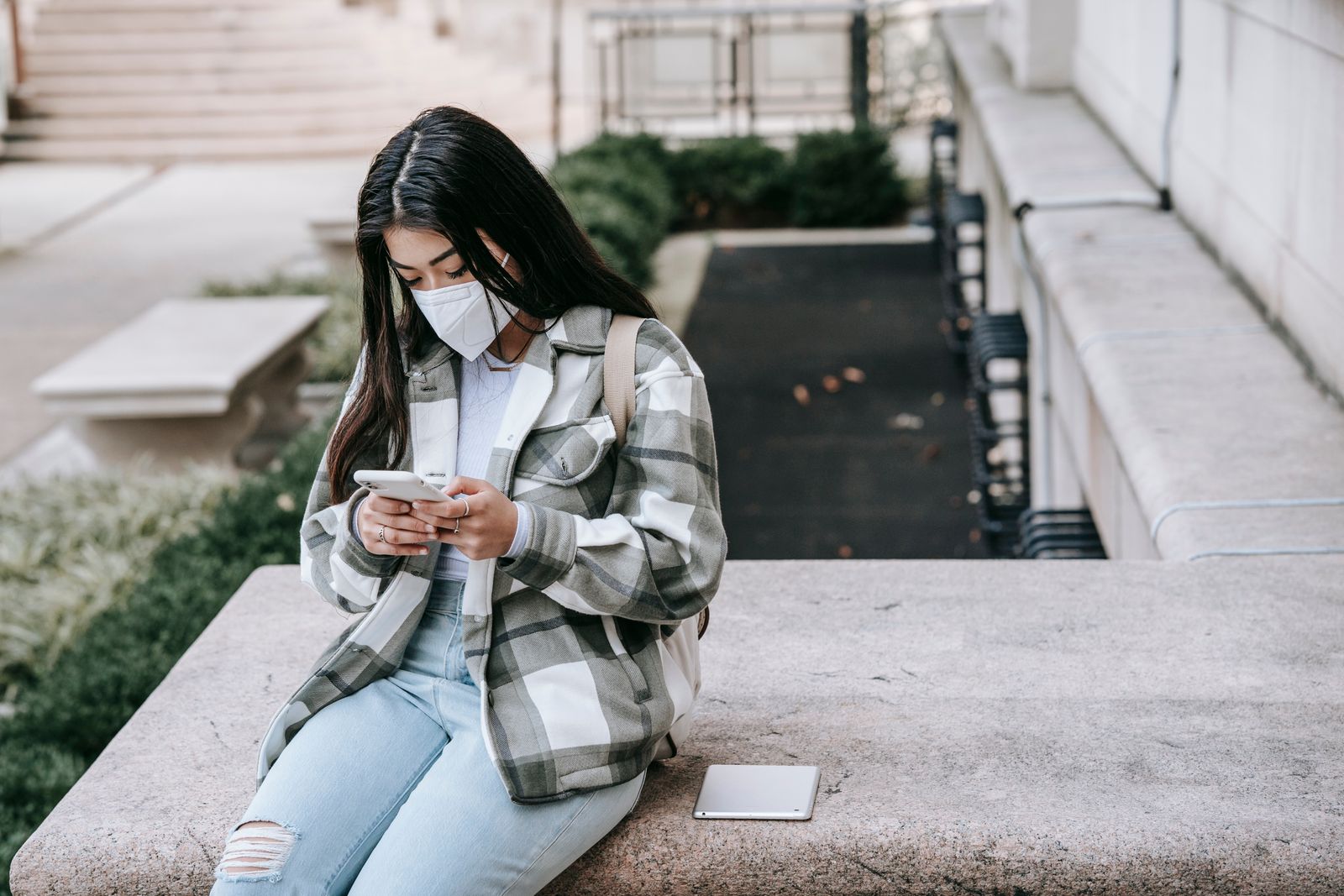 Faceless young female in casual clothes and mask sitting on stone fence while texting on cellphone near tablet in daylight
