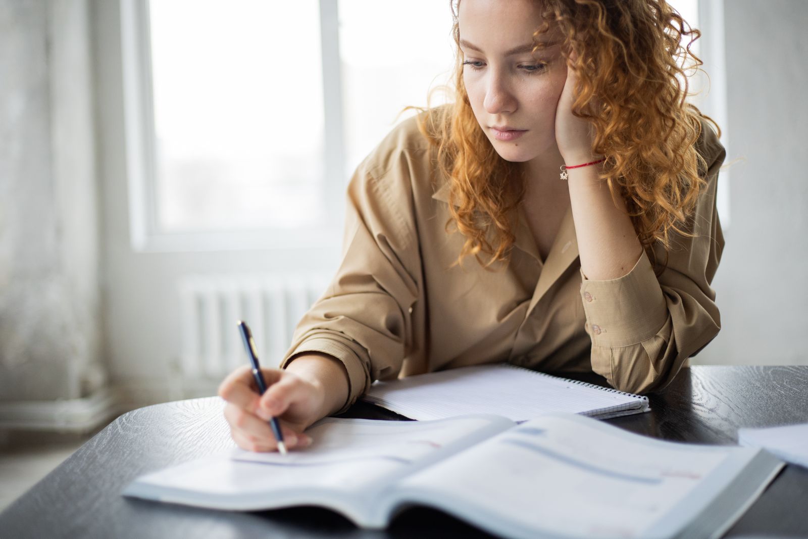 Diligent female student with copybook leaning on hand and taking notes in textbook at wooden table while studying in room