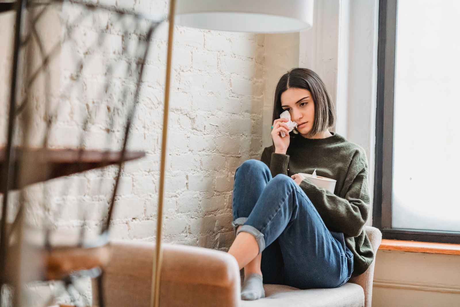 Full body of pensive young ethnic lady in casual clothes wiping tears with tissue while sitting on couch near window at home