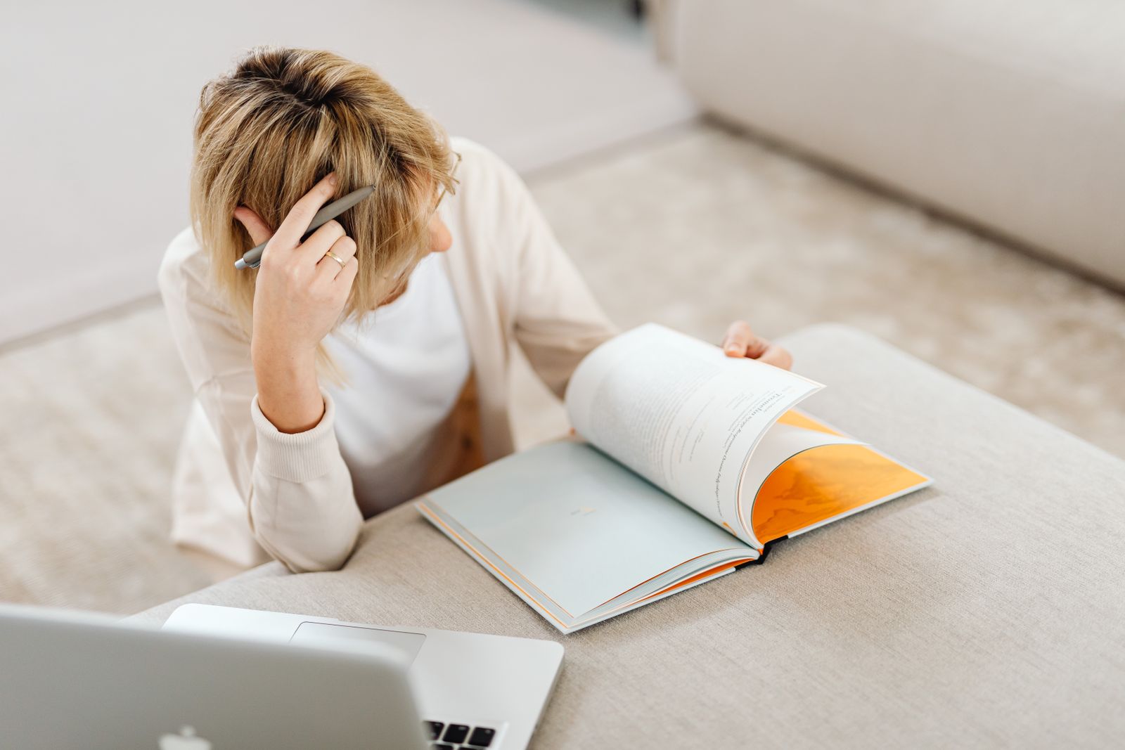 Blonde woman studying in living room with a book and laptop, creating a relaxed atmosphere.