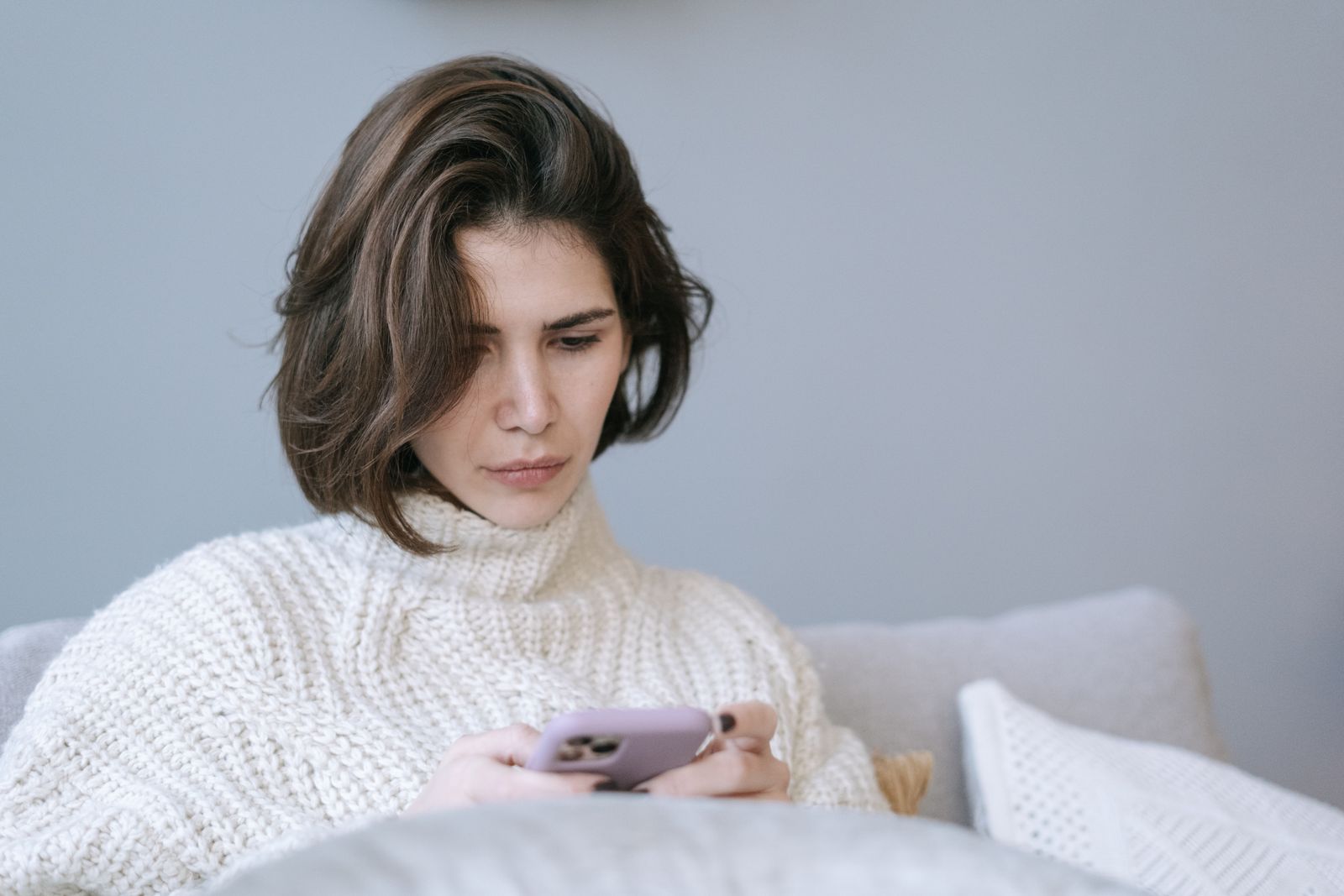 Woman in cozy knit sweater using a smartphone indoors, expressing a thoughtful mood.