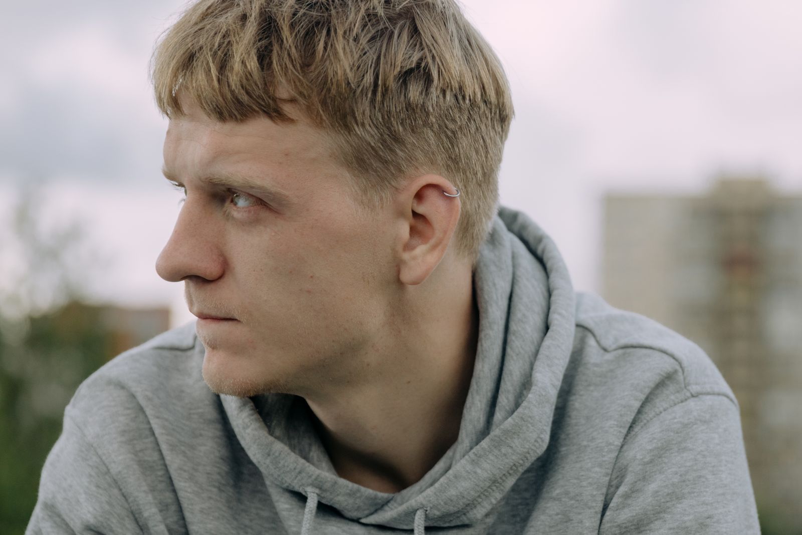 A young man in a gray hoodie looking thoughtfully into the distance outdoors.