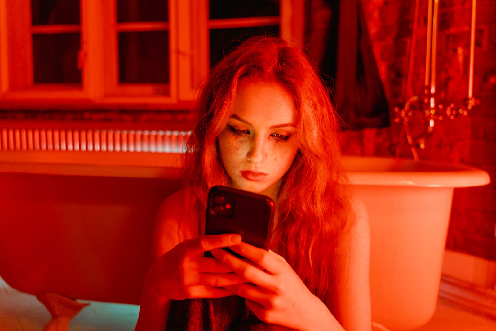 A young woman using a smartphone with a serious look in a red-lit bathroom.
