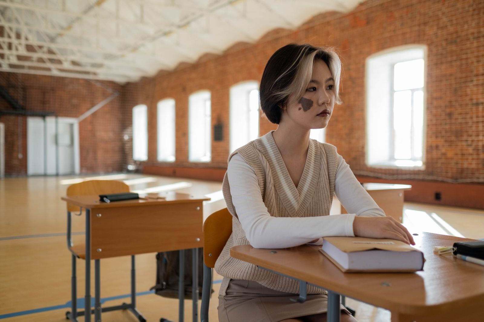 Young woman with birthmark sitting at a desk in a sunlit classroom with exposed brick walls.