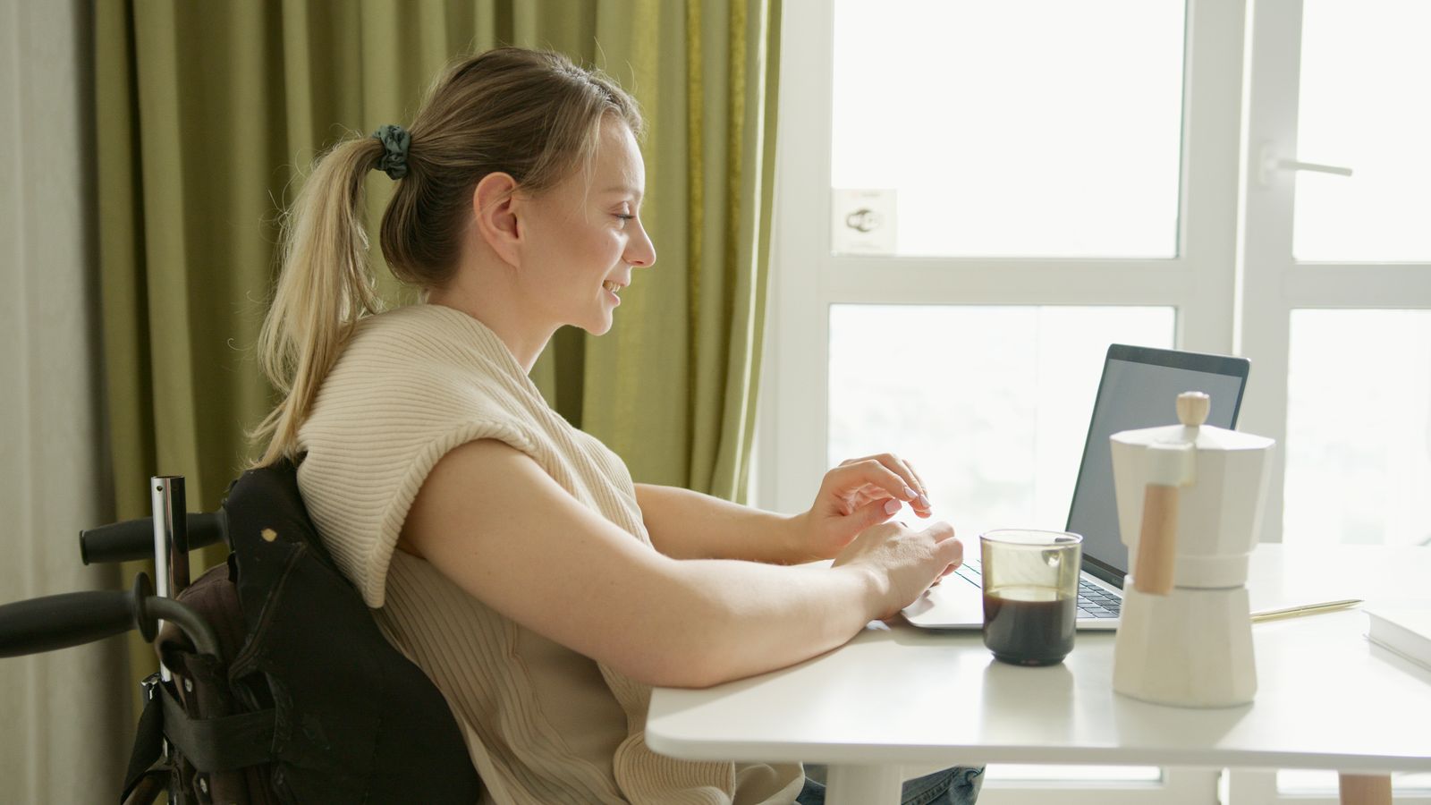 A woman in a wheelchair working on a laptop with coffee on the table, indoors.