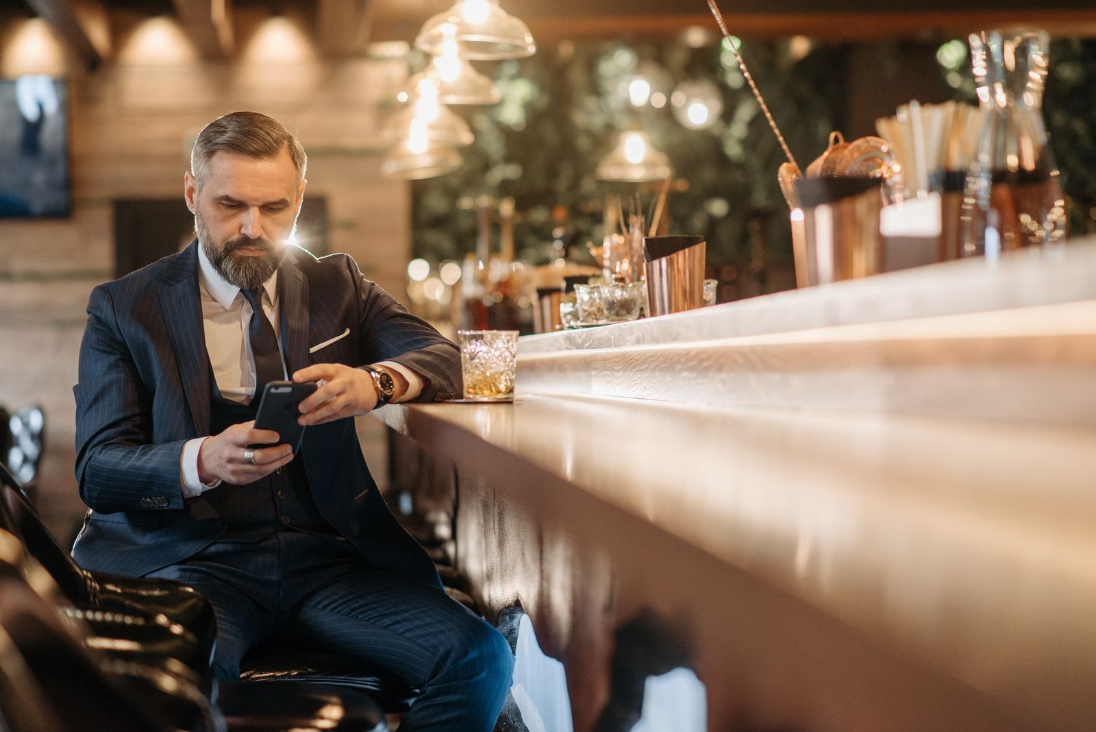 A businessman in a suit checks his smartphone while seated at a bar enjoying a whiskey.