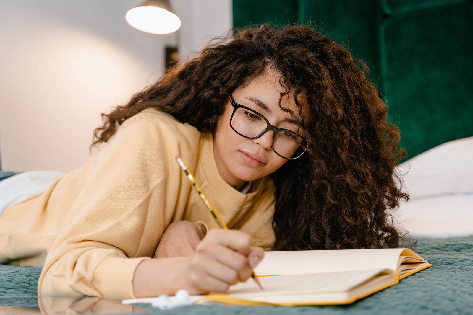 A young woman with glasses and curly hair studies on her bed, focusing on her notes.