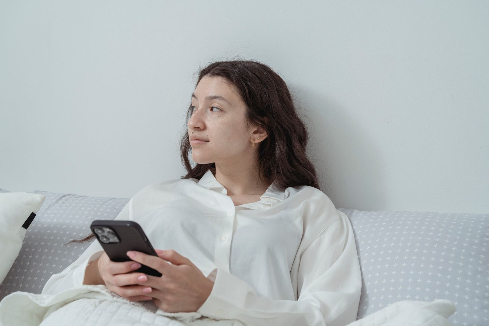 A woman in a white shirt sits on a bed, holding a smartphone, looking thoughtful.